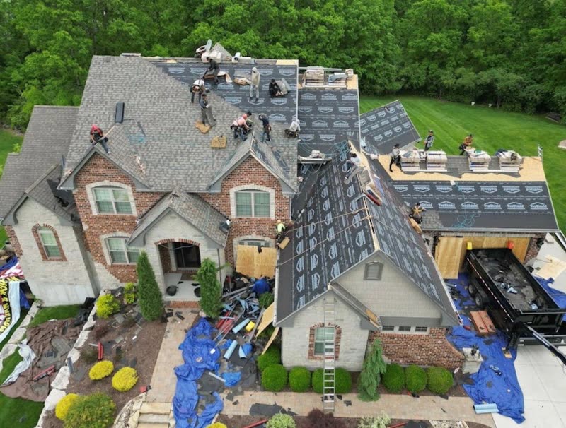 Roofing crew tearing off a brick and stone estate, ground protected with tarps and a debris trailer alongside